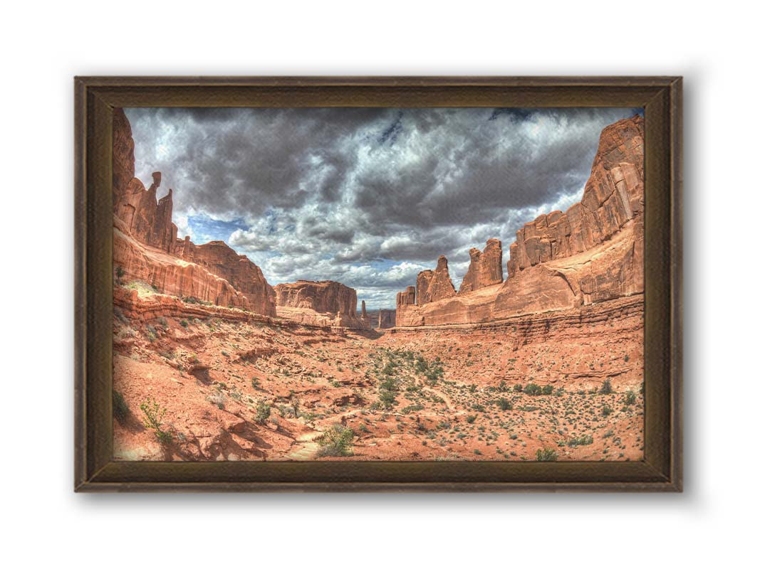 A photo of a tan, sandy, rocky valley along the Park Avenue hiking trail at Arches National Park in Utah. Gray and white clouds fill the sky. Printed on canvas and framed.