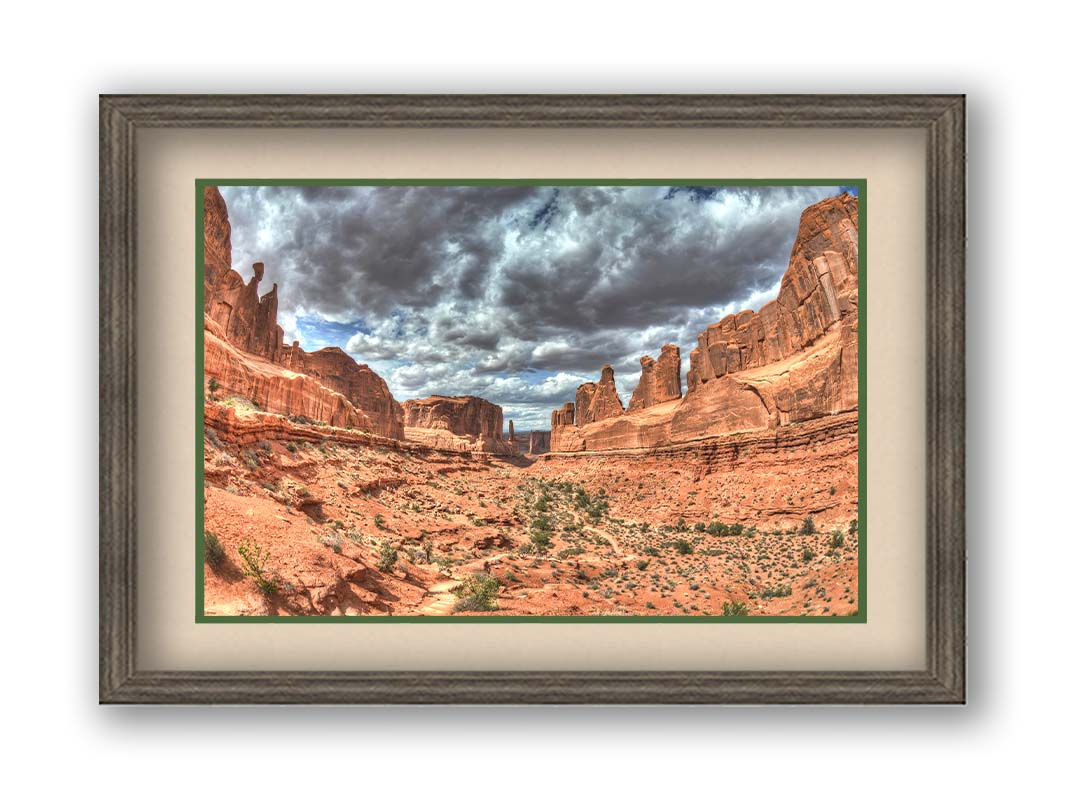 A photo of a tan, sandy, rocky valley along the Park Avenue hiking trail at Arches National Park in Utah. Gray and white clouds fill the sky. Printed on paper, matted, and framed.