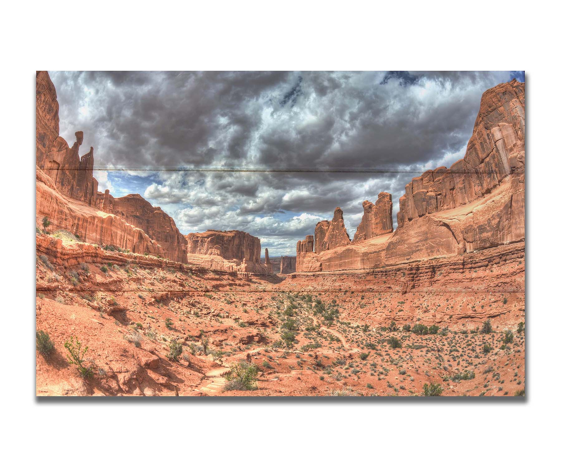 A photo of a tan, sandy, rocky valley along the Park Avenue hiking trail at Arches National Park in Utah. Gray and white clouds fill the sky. Printed on a box board.