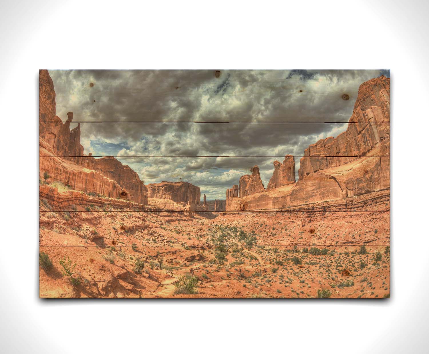 A photo of a tan, sandy, rocky valley along the Park Avenue hiking trail at Arches National Park in Utah. Gray and white clouds fill the sky. Printed on a wood pallet.