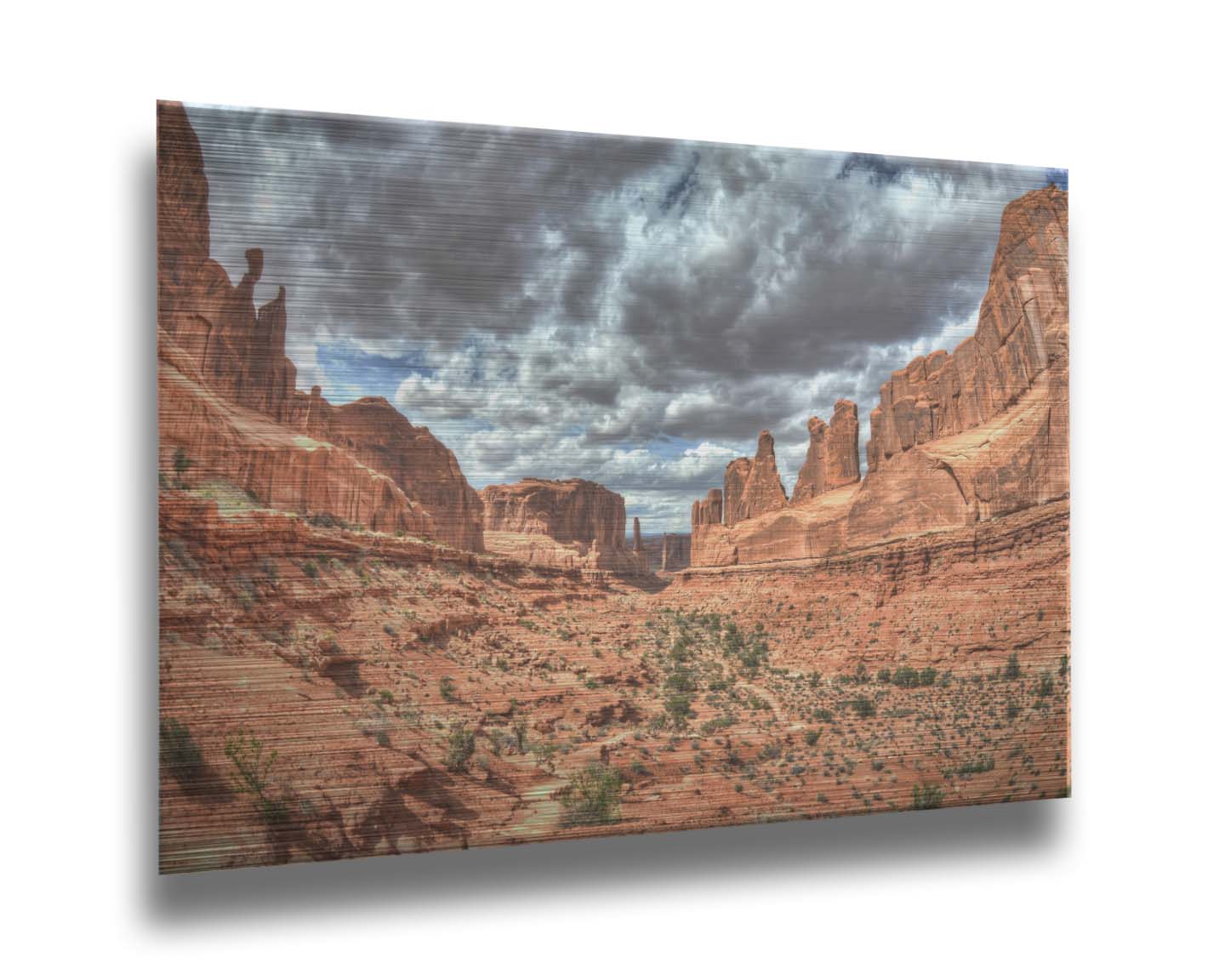 A photo of a tan, sandy, rocky valley along the Park Avenue hiking trail at Arches National Park in Utah. Gray and white clouds fill the sky. Printed on metal.