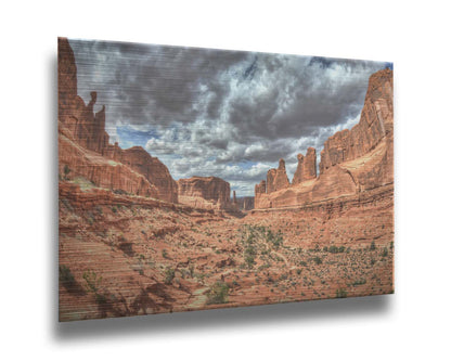 A photo of a tan, sandy, rocky valley along the Park Avenue hiking trail at Arches National Park in Utah. Gray and white clouds fill the sky. Printed on metal.