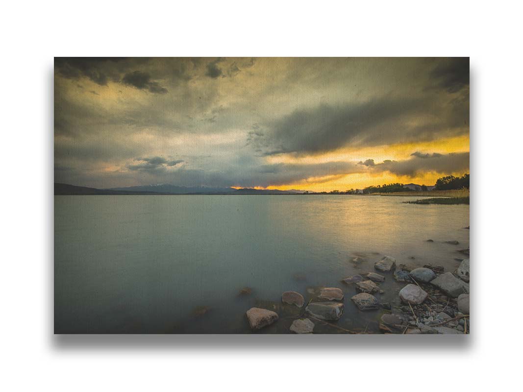 Photo of the sunset over Utah Lake at the American Fork Harbor. The yellow sky peeks through gray clouds over the calm, murky grays and blues of the lake. Printed on canvas.