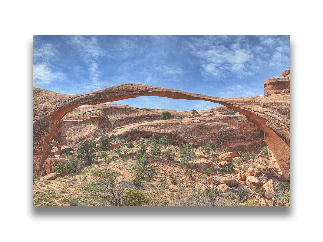 A photo of Landscape Arch at Arches National Park. A long, thin arch of orange and brown stone reaches across a bright blue sky. Printed on canvas.