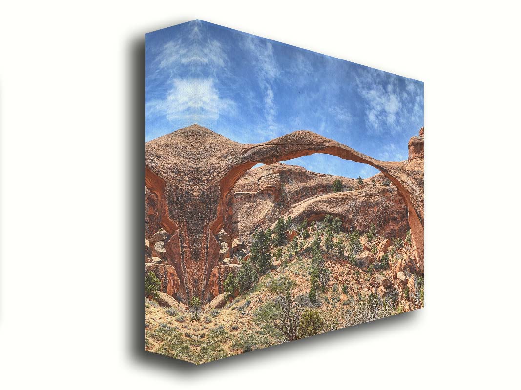 A photo of Landscape Arch at Arches National Park. A long, thin arch of orange and brown stone reaches across a bright blue sky. Printed on canvas.