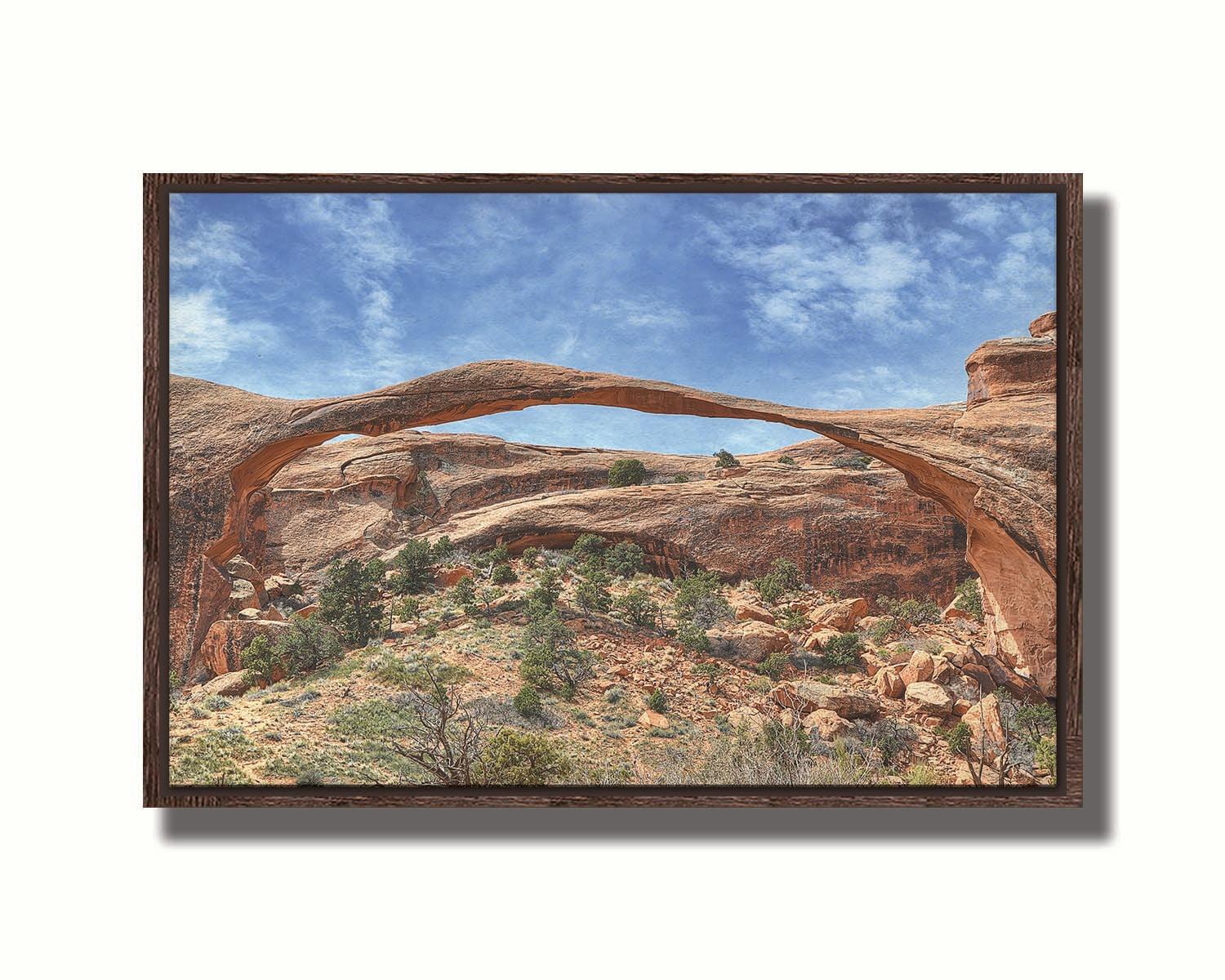 A photo of Landscape Arch at Arches National Park. A long, thin arch of orange and brown stone reaches across a bright blue sky. Printed on canvas in a float frame.