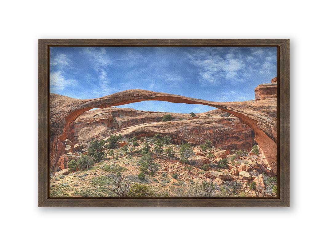 A photo of Landscape Arch at Arches National Park. A long, thin arch of orange and brown stone reaches across a bright blue sky. Printed on canvas and framed.