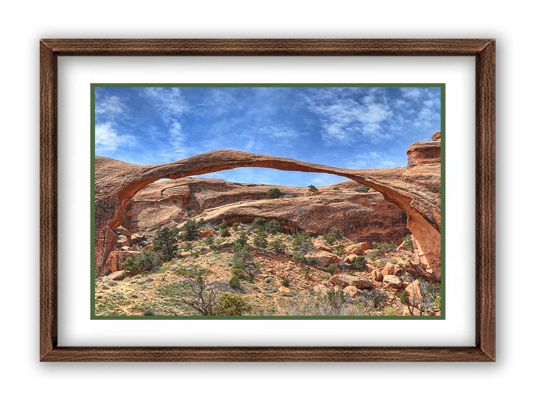 A photo of Landscape Arch at Arches National Park. A long, thin arch of orange and brown stone reaches across a bright blue sky. Printed on paper, matted, and framed.