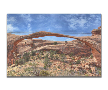 A photo of Landscape Arch at Arches National Park. A long, thin arch of orange and brown stone reaches across a bright blue sky. Printed on a box board.