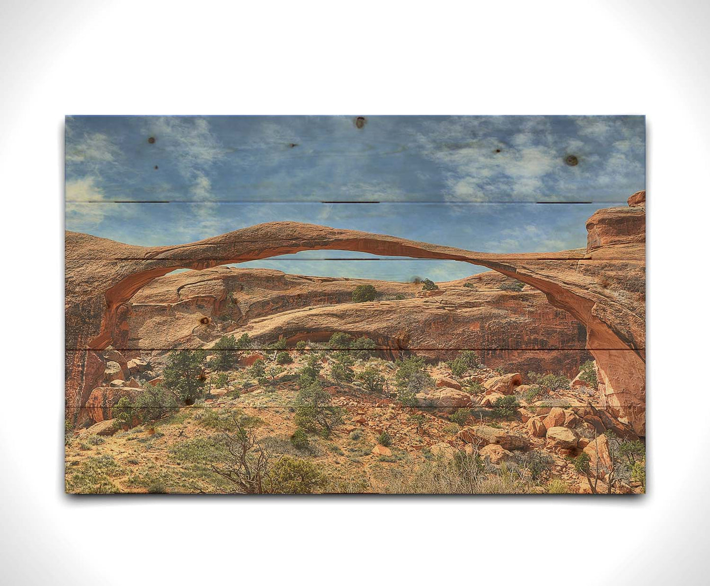 A photo of Landscape Arch at Arches National Park. A long, thin arch of orange and brown stone reaches across a bright blue sky. Printed on a wood pallet.