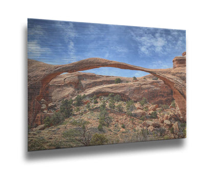 A photo of Landscape Arch at Arches National Park. A long, thin arch of orange and brown stone reaches across a bright blue sky. Printed on metal.