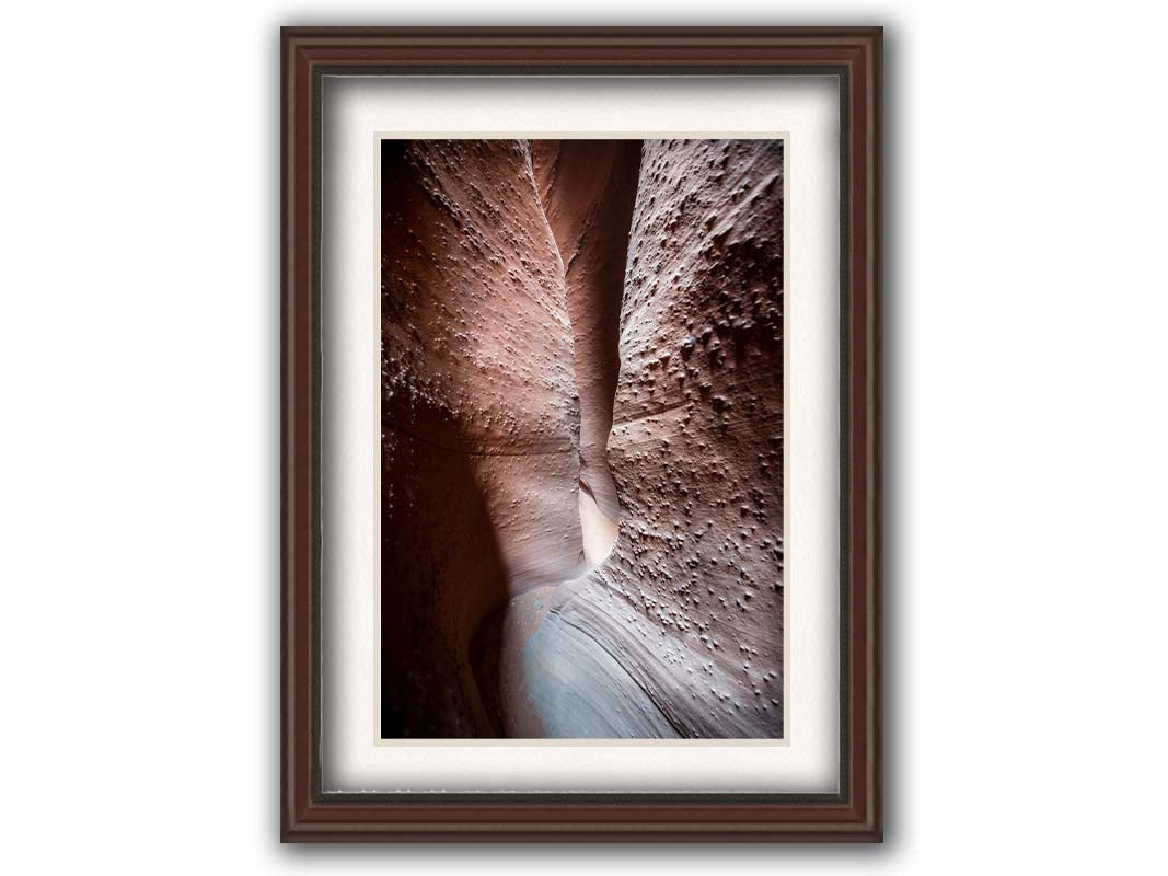 A photo of the slot canyon called Spooky near Escalante, Utah. It features a narrow crevice of lrough and smooth layers of stone. Printed on paper, matted, and framed.