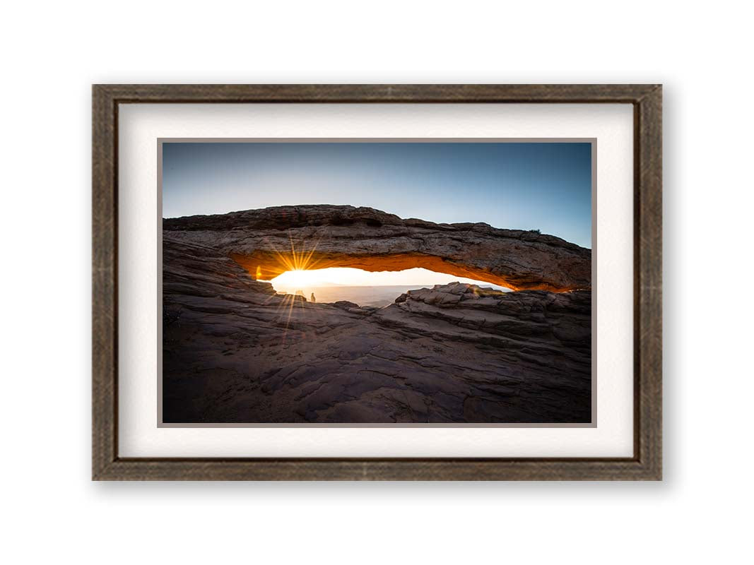 Photo of a stone arch from a national park in Utah. The sunrise site just between the horizon and the underside of the arch, creating a beautiful orange highlight to the underside of the stone structure. Printed on paper, matted, and framed.