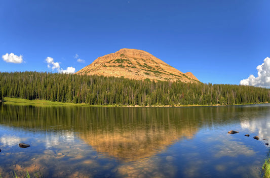 Photo of Mirror Lake in Utah. A mountain, forest, blue sky, and fluffy clouds are reflected on the lake water.
