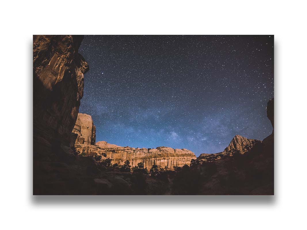 A photo of a clear, starry night sky from the Capitol Reef National Park. The stony plateaus encircle the view of the stars. Printed on canvas.