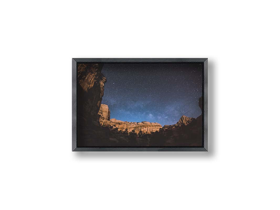 A photo of a clear, starry night sky from the Capitol Reef National Park. The stony plateaus encircle the view of the stars. Printed on canvas and framed.