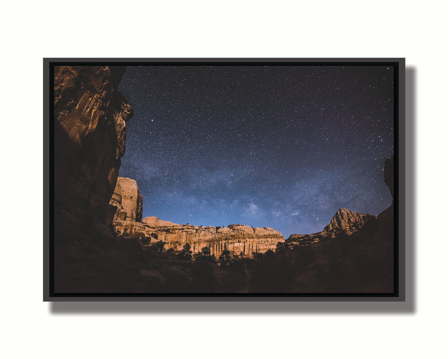 A photo of a clear, starry night sky from the Capitol Reef National Park. The stony plateaus encircle the view of the stars. Printed on canvas in a float frame.