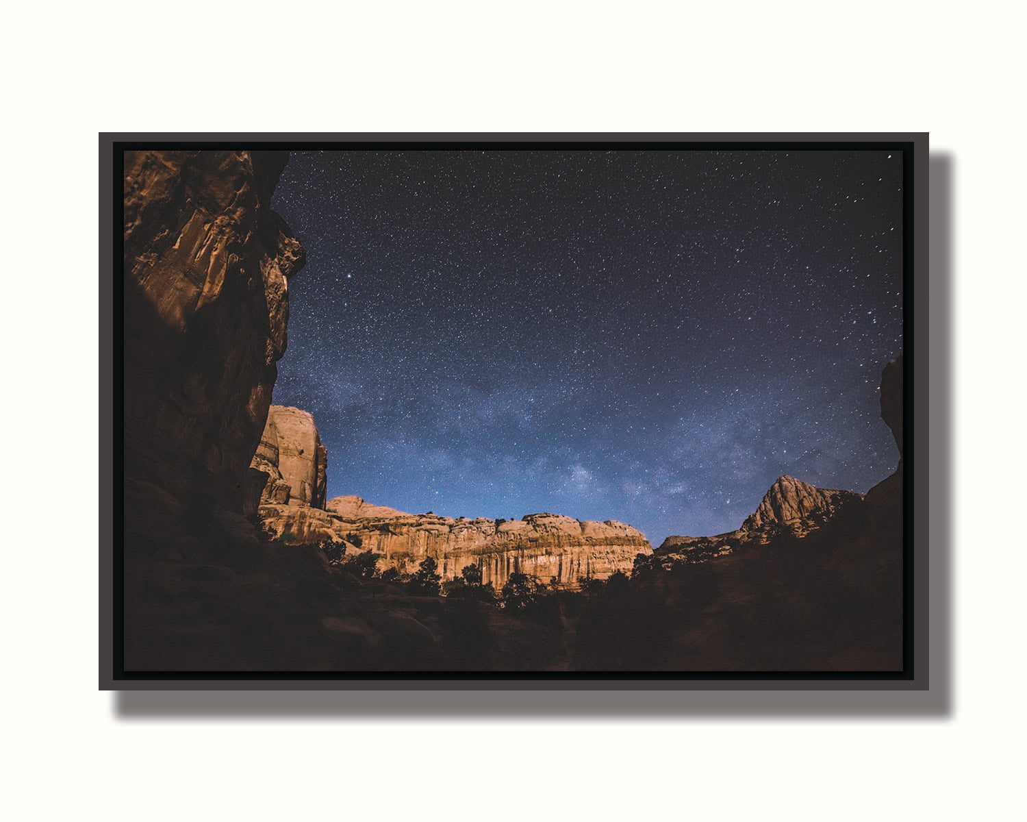 A photo of a clear, starry night sky from the Capitol Reef National Park. The stony plateaus encircle the view of the stars. Printed on canvas in a float frame.