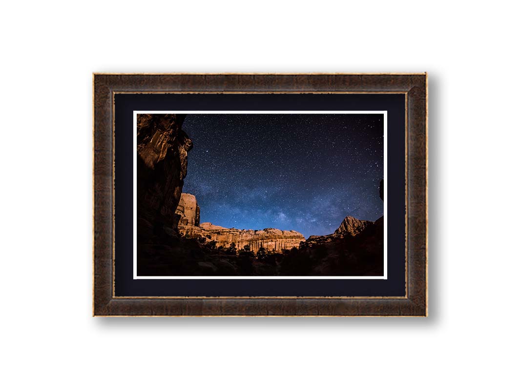 A photo of a clear, starry night sky from the Capitol Reef National Park. The stony plateaus encircle the view of the stars. Printed on paper, matted, and framed.