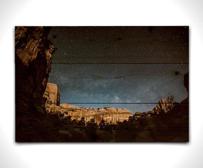 A photo of a clear, starry night sky from the Capitol Reef National Park. The stony plateaus encircle the view of the stars. Printed on a wood pallet.