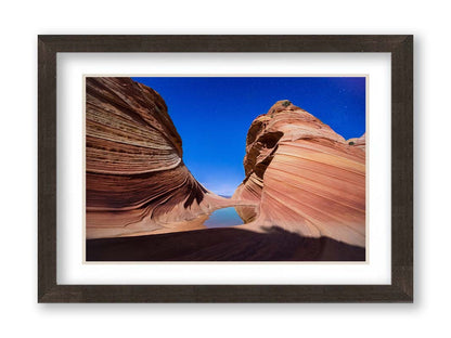 Photo of the "Wave" rock formation at Coyote Buttes North in Utah. A bright night shot captures stripes of sandstone layers forming orange waves against a starry blue sky. Printed on paper, matted, and framed.