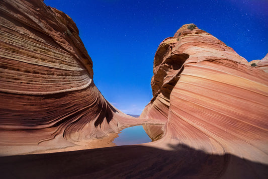 Photo of the "Wave" rock formation at Coyote Buttes North in Utah. A bright night shot captures stripes of sandstone layers forming orange waves against a starry blue sky.