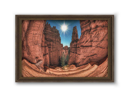 Photo of the Navajo Loop Switchbacks rock formation at Bryce Canyon National Park in Utah. It is taken wit a fish eye lens perspective which emphasizes the height and depth of the structures. A bright blue sky peeks out between parallel walls of red stone, and the bright white rays of the perfectly centered sun shine down between. Printed on canvas and framed.