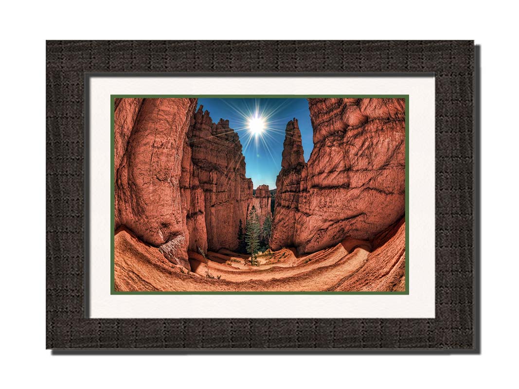 Photo of the Navajo Loop Switchbacks rock formation at Bryce Canyon National Park in Utah. It is taken wit a fish eye lens perspective which emphasizes the height and depth of the structures. A bright blue sky peeks out between parallel walls of red stone, and the bright white rays of the perfectly centered sun shine down between. Printed on paper, matted, and framed.