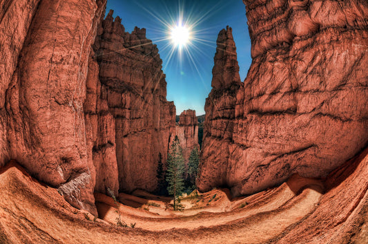 Photo of the Navajo Loop Switchbacks rock formation at Bryce Canyon National Park in Utah. It is taken with a fish eye lens perspective, which emphasizes the height and depth of the structures. A bright blue sky peeks out between parallel walls of red stone, and the bright white rays of the perfectly centered sun shine down between. 