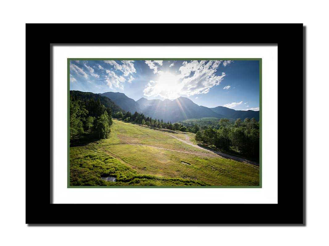 A photo of the sun shining brightly over Mount Timpanogos. The hillsides and forest are the fresh green of springtime. Printed on paper, matted, and framed.