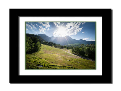 A photo of the sun shining brightly over Mount Timpanogos. The hillsides and forest are the fresh green of springtime. Printed on paper, matted, and framed.