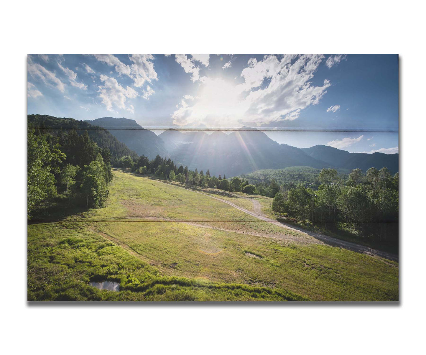 A photo of the sun shining brightly over Mount Timpanogos. The hillsides and forest are the fresh green of springtime. Printed on a box board.