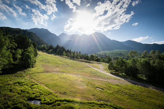 A photo of the sun shining brightly over Mount Timpanogos. The hillsides and forest are the fresh green of springtime. 