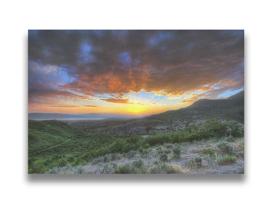 A photo of the sunset at Piestewa Peak in Provo Canyon, Utah. The gold light from the sun reflects off gray clouds about the green foliage surrounding the mountain. Printed on canvas.