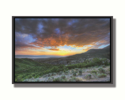 A photo of the sunset at Piestewa Peak in Provo Canyon, Utah. The gold light from the sun reflects off gray clouds about the green foliage surrounding the mountain. Printed on canvas in a float frame.