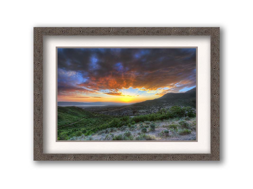 A photo of the sunset at Piestewa Peak in Provo Canyon, Utah. The gold light from the sun reflects off gray clouds about the green foliage surrounding the mountain. Printed on paper, matted, and framed.