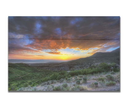 A photo of the sunset at Piestewa Peak in Provo Canyon, Utah. The gold light from the sun reflects off gray clouds about the green foliage surrounding the mountain. Printed on a box board.