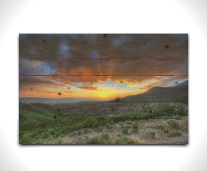 A photo of the sunset at Piestewa Peak in Provo Canyon, Utah. The gold light from the sun reflects off gray clouds about the green foliage surrounding the mountain. Printed on a wood pallet.