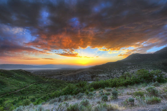 A photo of the sunset at Piestewa Peak in Provo Canyon, Utah. The gold light from the sun reflects off gray clouds about the green foliage surrounding the mountain.