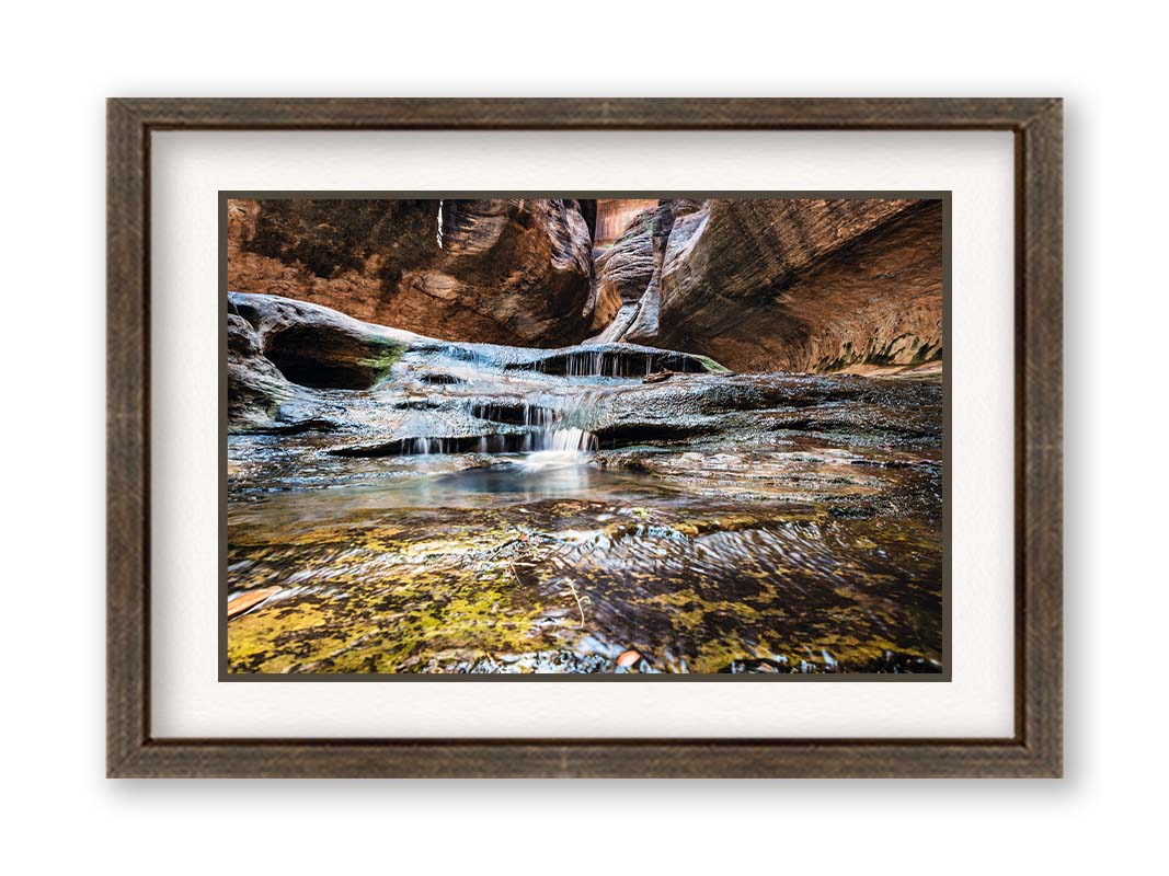 A photo of the Left Fork North Creek flowing through the Subway slot canyon in Utah's Zion National Park. It flows toward the camera, tumbling over slabs in the sandstone to create small waterfalls. Printed on paper, matted, and framed.