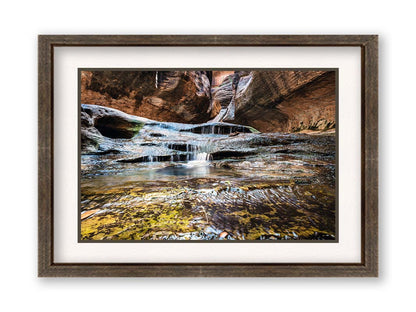 A photo of the Left Fork North Creek flowing through the Subway slot canyon in Utah's Zion National Park. It flows toward the camera, tumbling over slabs in the sandstone to create small waterfalls. Printed on paper, matted, and framed.