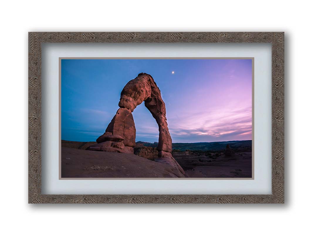 A photo of the Delicate Arch rock formation in Arches National Park, Utah. The blues and purples of the sky at sunset compliment the oranges and browns of the sandstone arch. Printed on paper, matted, and framed.