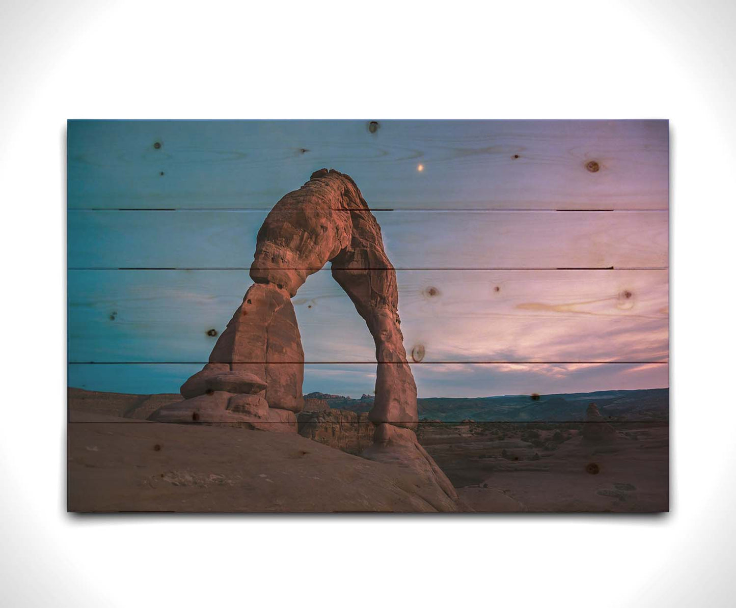 A photo of the Delicate Arch rock formation in Arches National Park, Utah. The blues and purples of the sky at sunset compliment the oranges and browns of the sandstone arch. Printed on a wood pallet.