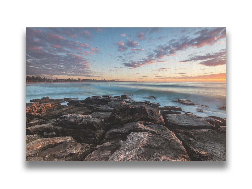 A photo of the sunrise at Manly Beach in Sydney, Australia. The city sits in the background as waves roll against the rocks. Printed on canvas.