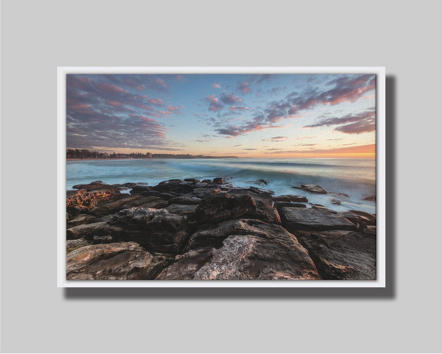 A photo of the sunrise at Manly Beach in Sydney, Australia. The city sits in the background as waves roll against the rocks. Printed on canvas in a float frame.