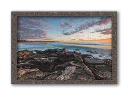 A photo of the sunrise at Manly Beach in Sydney, Australia. The city sits in the background as waves roll against the rocks. Printed on canvas and framed.