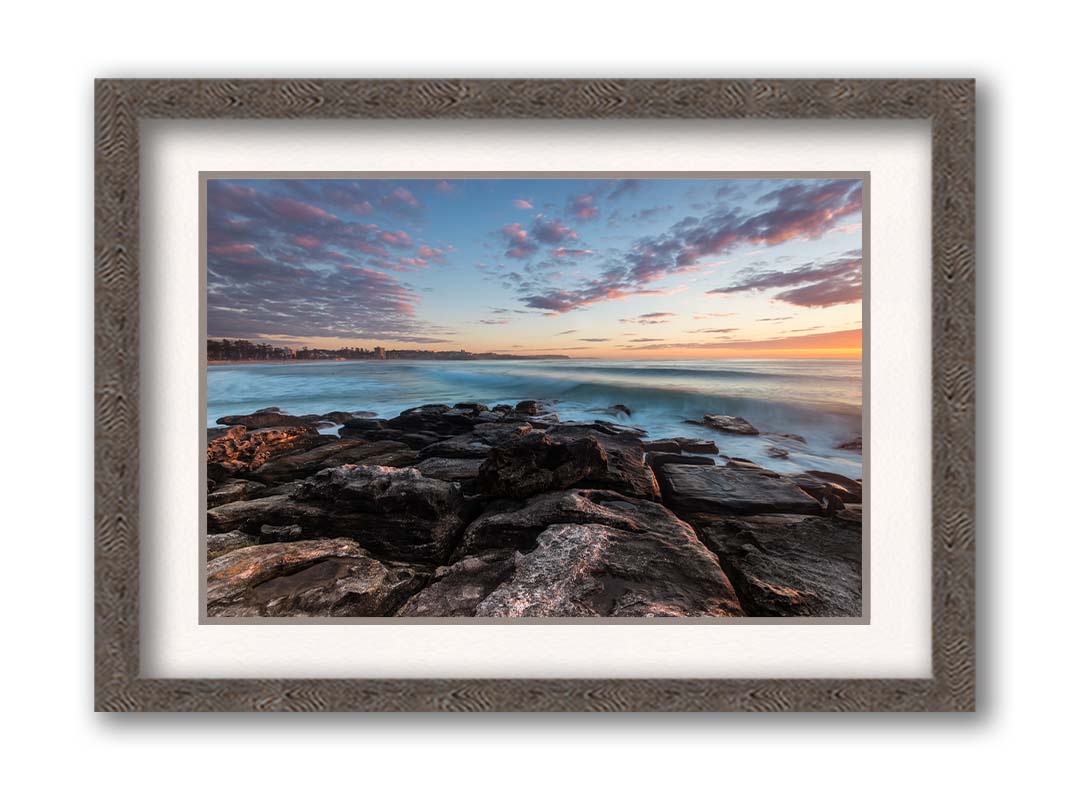 A photo of the sunrise at Manly Beach in Sydney, Australia. The city sits in the background as waves roll against the rocks. Printed on paper, matted, and framed.