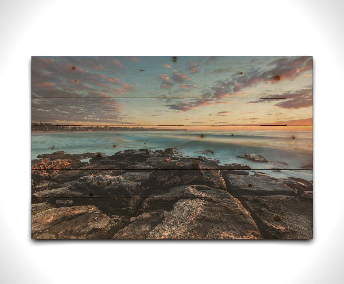 A photo of the sunrise at Manly Beach in Sydney, Australia. The city sits in the background as waves roll against the rocks. Printed on a wood pallet.