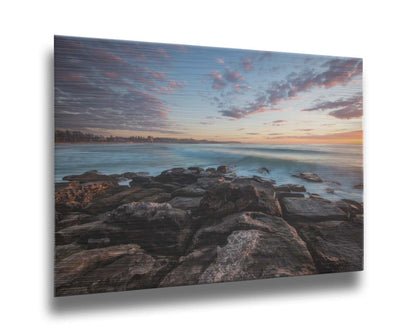 A photo of the sunrise at Manly Beach in Sydney, Australia. The city sits in the background as waves roll against the rocks. Printed on metal.