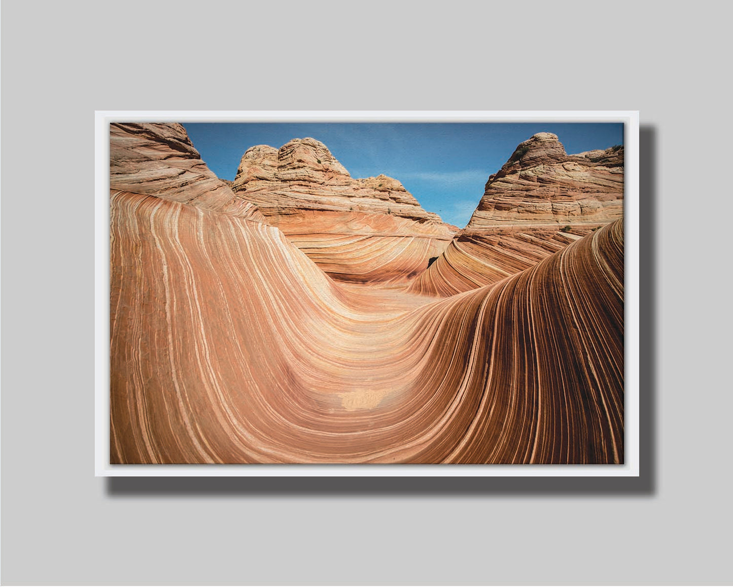 A photo of the "Wave" rock formation at Coyote Buttes North in Utah. The wavy sandstone layers create rolling waves where they have been carved out of the mountainside. The orange hues of the stone are sharply contrasted by the vibrant blue of the sky. Printed on canvas in a float frame.